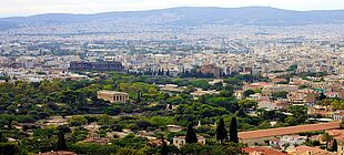 Es ist die Stadt Athen von Weitem zu sehen. Im Hintergrund sind viele Häuser und im Vordergrund etwas Natur.