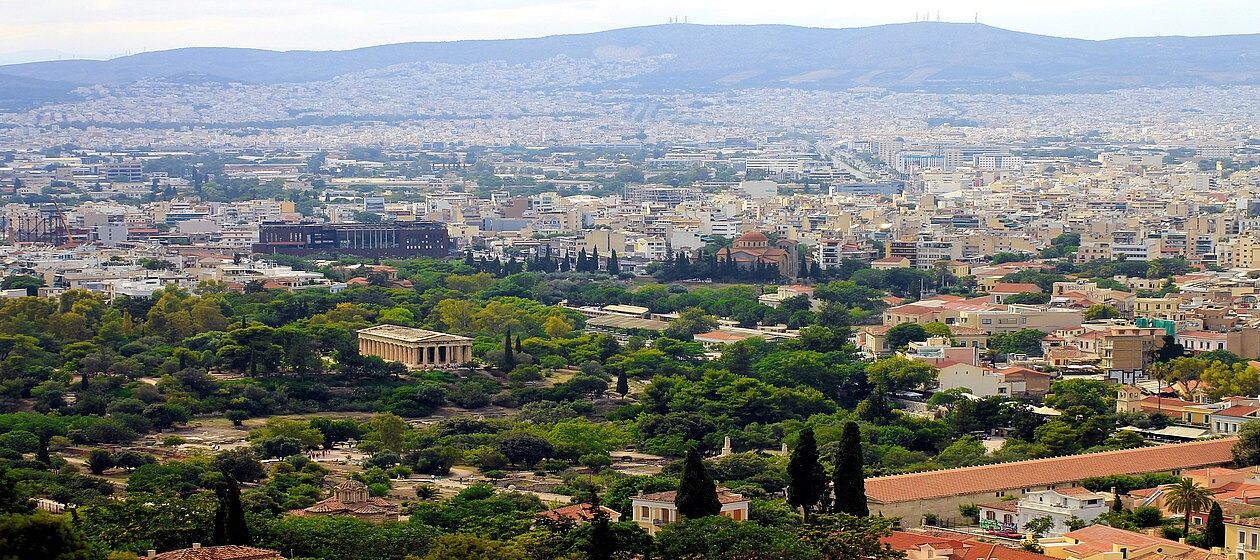 Es ist die Stadt Athen von Weitem zu sehen. Im Hintergrund sind viele Häuser und im Vordergrund etwas Natur.