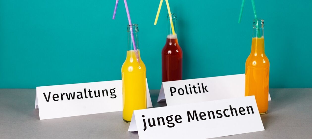 Drei Tischkarten mit den Aufschriften "Verwaltung", "Politik" und "junge Menschen" neben drei bunten Limo-Flaschen