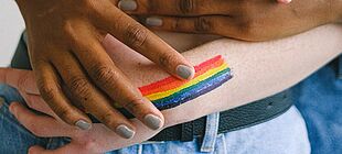 Zwei Personen unterschiedlicher Hautfarbe mit umeinander gelegten Armen, auf den Arm der einen Person ist eine Regenbogenflagge gemalt