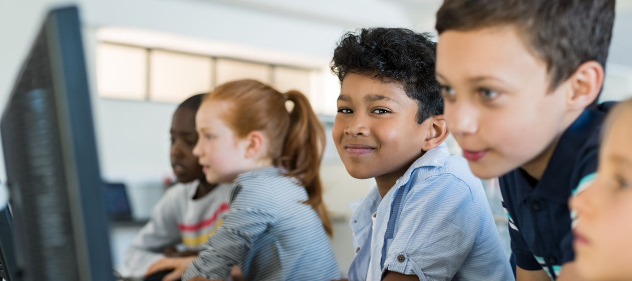 Kinder unterschiedlicher Herkunft sitzen vor Computern in der Schule und lächeln.