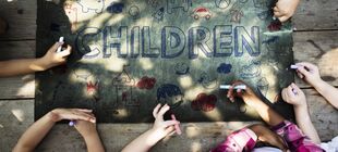 Kindern halten Kreidestifte in den Händen und sitzen um einem Tisch, auf dem ein Plakat mit der Auschrift Children liegt