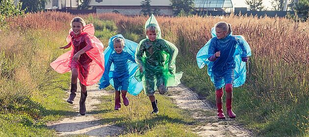 Vier Kinder mit bunten Regenponchos und Gummistiefeln rennen einen Weg entlang