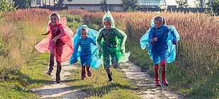 Vier Kinder mit bunten Regenponchos und Gummistiefeln rennen einen Weg entlang