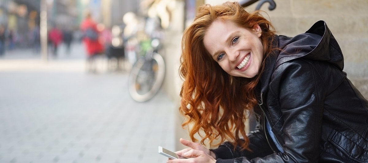 Eine lachende junge Frau sitzt auf einer Treppe in der Stadt mit ihrem Handy in der Hand