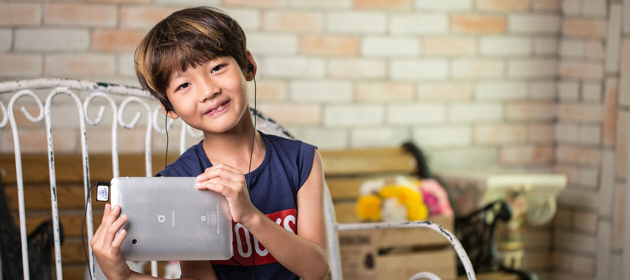 Ein Kind hält ein Tablet in den Händen, auf dem es etwas mit Kopfhörern hört.