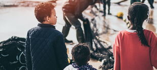 Drei Kinder mit dunkler Hautfarbe stehen am Meer, halten sich an den Händen, im Hintergrund sind weitere Personen zu sehen