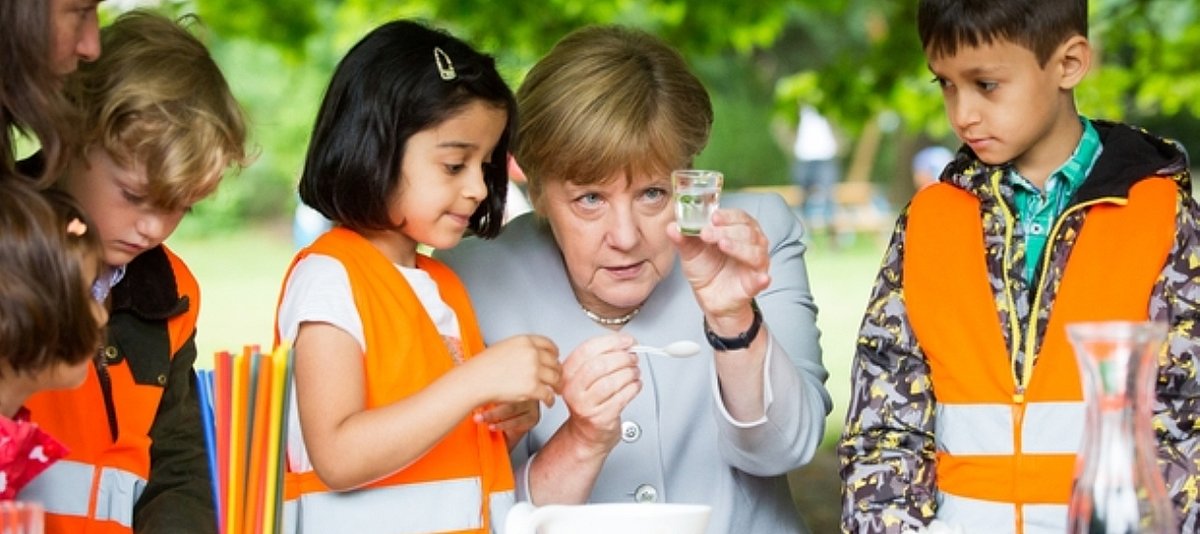Bundespräsidentin Angela Merkel sitzt im Freien zwischen Kita-Kindern und betrachtet ein Glas.