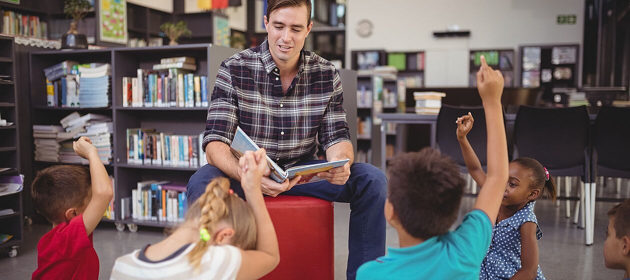 Eine erwachsene Person liest einer Gruppe Kindern aus einem Buch vor, die Kinder melden sich