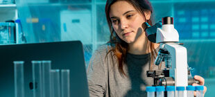 Eine junge Frau sitzt im Labor vor einem Mikroskop und schaut auf den Bildschirm eines Laptops.