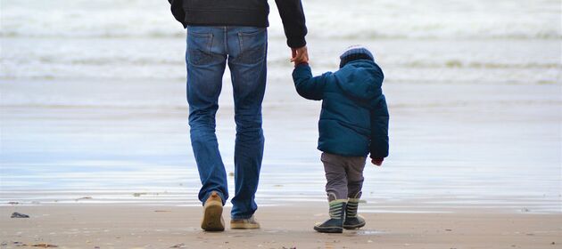 Mann hält Kleinkind mit Winterjacke an der Hand am Strand