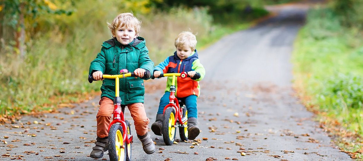 Zwei kleine Kinder sind auf einem Waldweg mit Laufrädern unterwegs