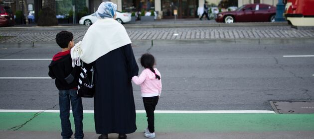 Eine Frau mit Kopftuch hält ihre beiden Kinder an der Hand und möchte über eine Straße gehen.