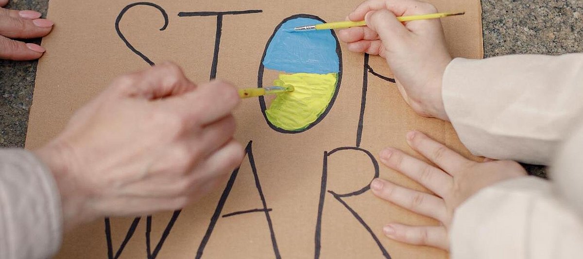 Zwei Menschen malen ein Plakat, auf dem Stop War steht, das O ist in den Farben der Ukraineflagge ausgemalt