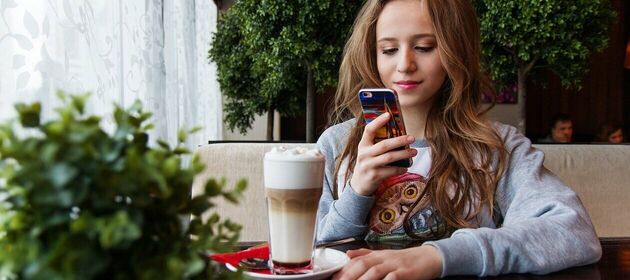 Ein Mädchen im Teenageralter sitzt vor einem Latte Macchiato in einem Café und schaut lächelnd auf das Smartphone in ihrer Hand. 