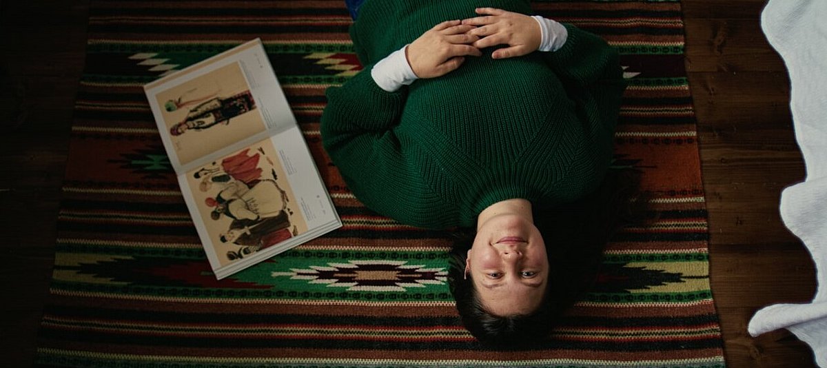 Das Bild zeigt eine Szene aus dem Dokumentarfilm „Generation Ukraine“, zu sehen ist eine junge Person, die auf dem Rücken liegt und lächelt, neben ihr liegt ein aufgeschlagenes Buch