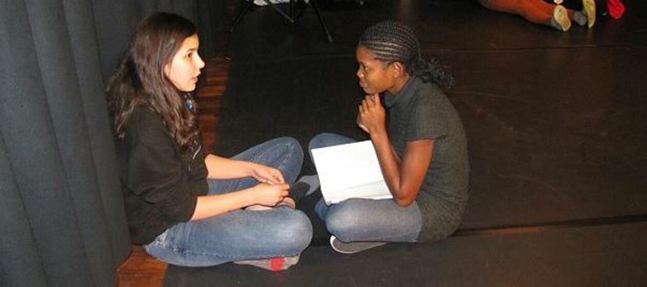 Ein europäisches und ein afrikanisches Mädchen sitzen im Schneidersitz auf dem Boden und üben gemeinsam den Text für ein Theaterstück.