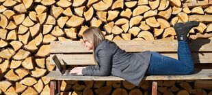 Eine junge Frau liegt auf einer sonnenbeschienenen Holzbank vor einer Holzscheitwand und arbeitet am Laptop.
