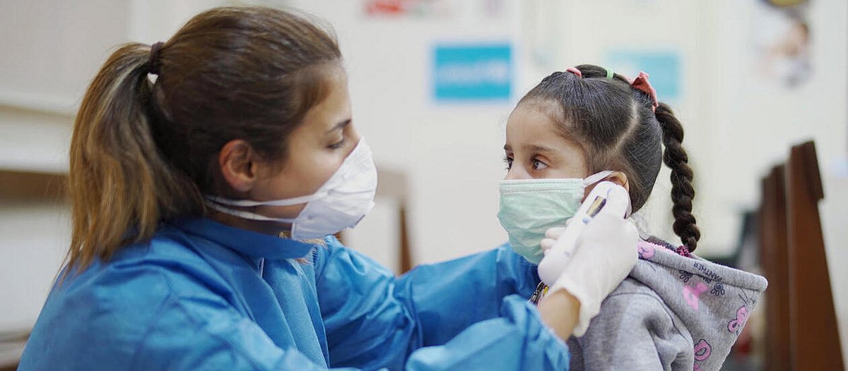 Eine Krankenschwester mit Mundschutz misst bei einem Mädchen Fieber.