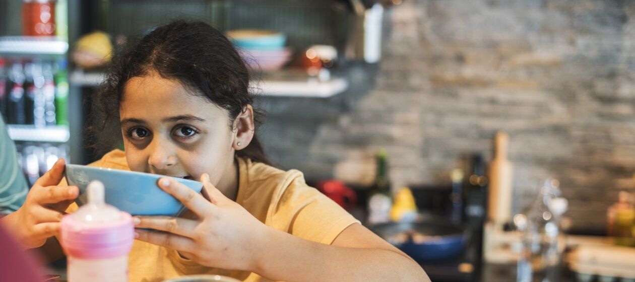 Ein Mädchen mit dunklen Haaren sitzt am Frühstückstisch und schaut ernst von ihrer Müslischale hoch