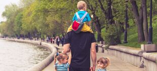 Eine erwachsene Person spaziert eine Promenade entland, an jeder Hand mit einem Kind, ein weiteres Kind sitzt auf den Schulter der Person