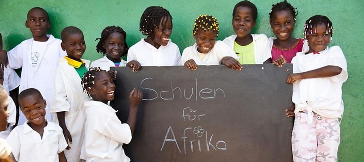 Schülerinnen und Schüler der Katepaschule in Angola stehen vor einer grünen Wand und halten eine Tafel mit der Aufschrift "Schulen für Afrika". 