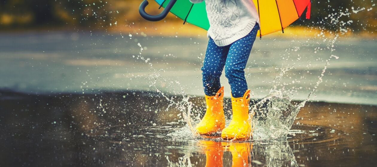 Ein Kind mit buntem Regenschirm in der Hand springt mit Gummistiefeln in eine Pfütze