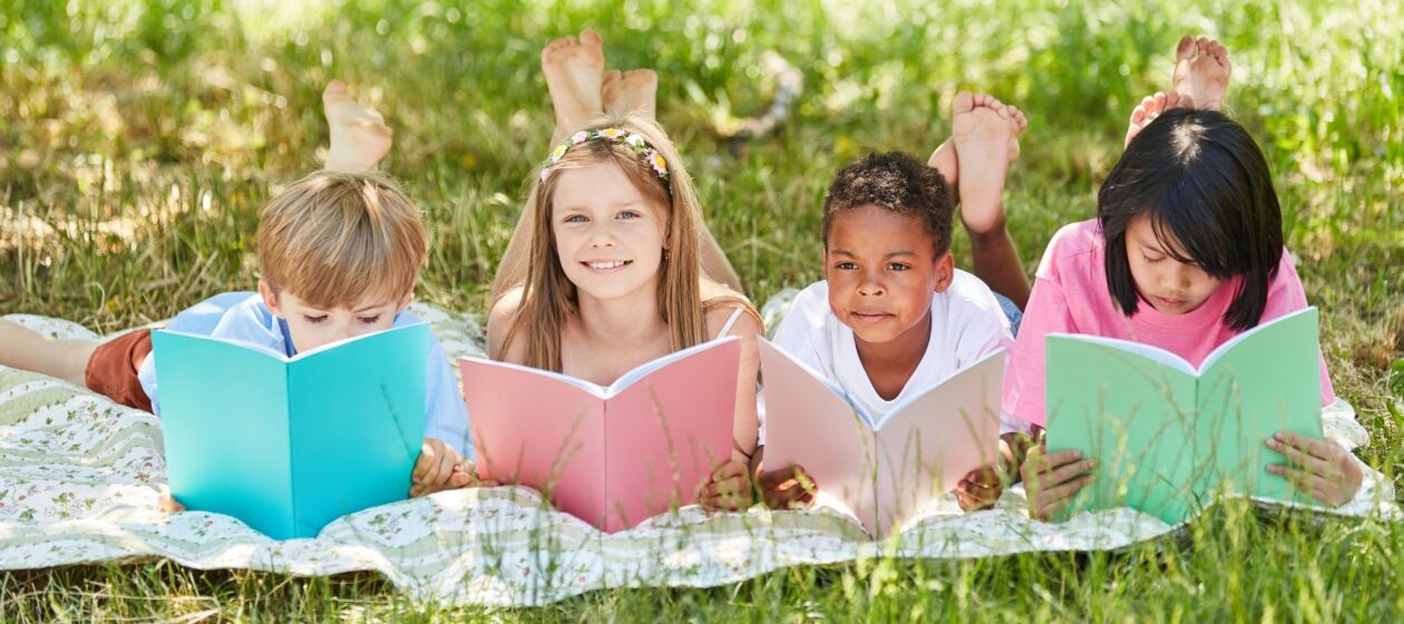 Vier Grundschulkinder liegen auf einer Wiese und lesen in unterschiedlich farbigen Schulheften