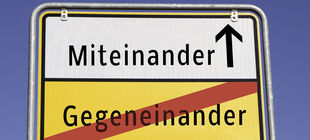 Ein Straßenschild zeigt durchgestrichen das Wort Gegeneinander und mit Richtungspfeil das Wort Miteinander.