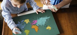 Ein Junge malt mit Kreide, zu sehen ist ein Bild der Weltkarte, im Hintergrund steht eine erwachsene Person und hält Kreidestücke