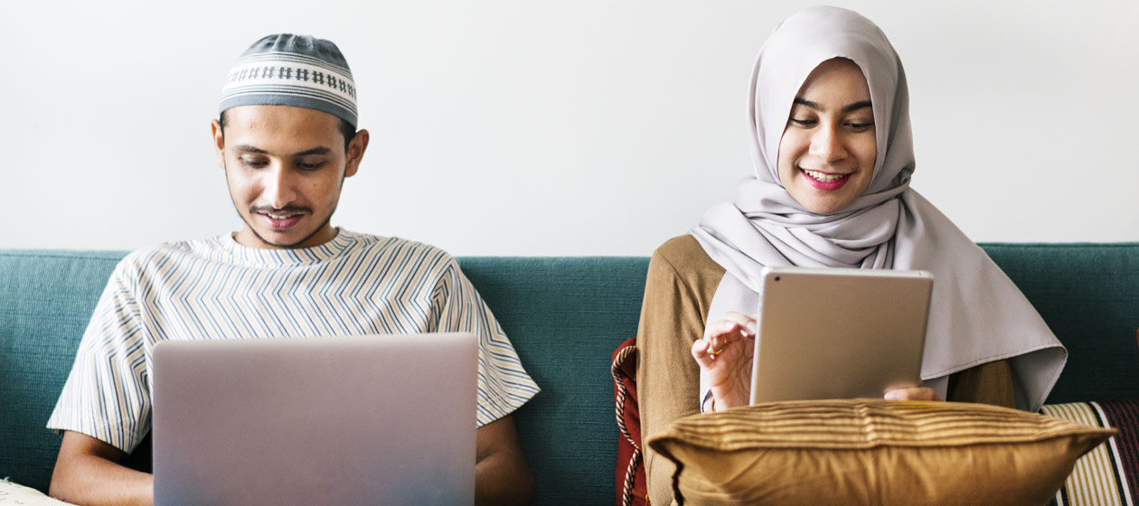 Zwei junge, muslimische Menschen sitzen auf einem Sofa mit Laptop und Tablet und lächeln