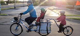 Eine erwachsene Person und zwei Kinder fahren mit Fahrradhelmen auf einem Tridem
