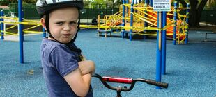 Ein Junge mit saurem Gesichtsausdruck hockt auf seinem BMX-Rad vor einem abgesperrten Spielplatz.