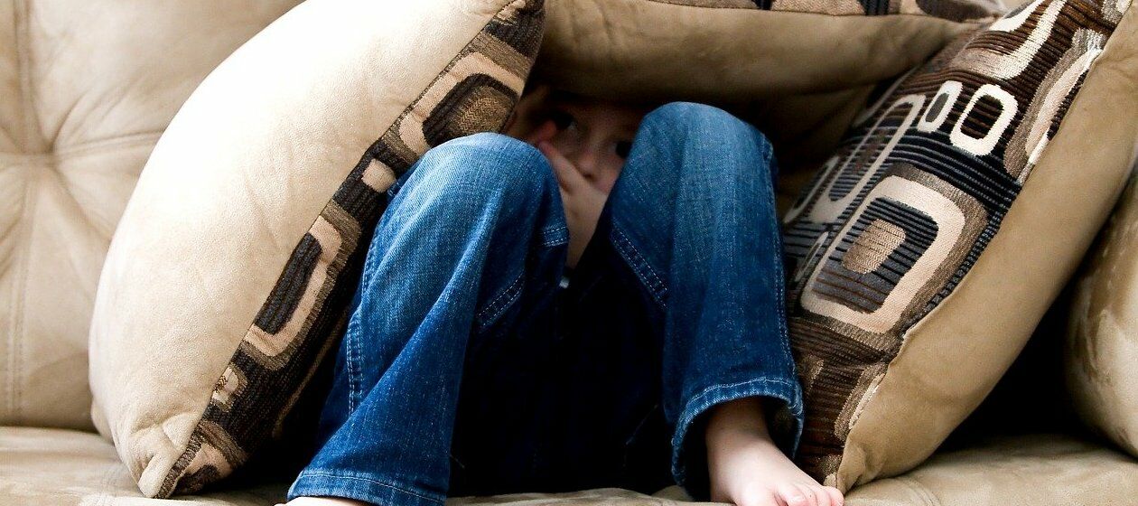 Ein Junge in blauen Jeans blickt aus seinem Kissenversteck auf einem Sofa in die Kamera.