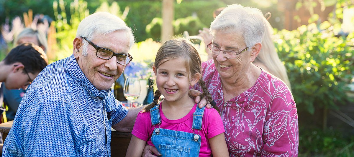 Großeltern sitzen mit ihrer Enkelin im Garten und lachen in die Kamera. 
