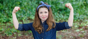 Ein Mädchen im Grundschulalter steht vor einer Hecke und zeigt mit erhobenen Fäusten eine Geste der Stärke und lächelt dabei.