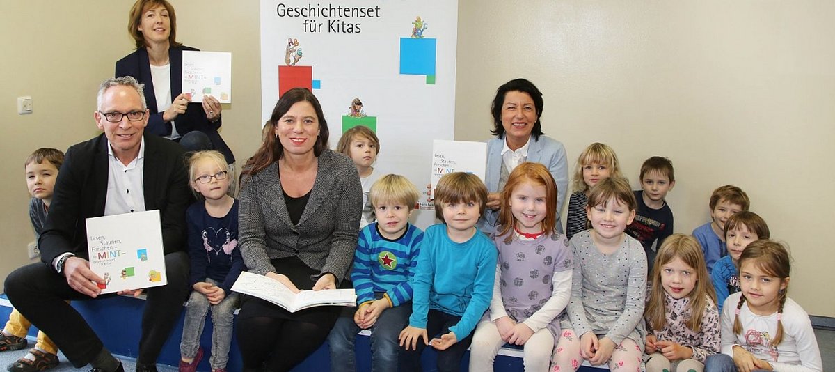 Kinder des Berliner Kindergartens Veteranenstraße sitzen neben Frau Scheeres und den anderen Beteiligten in einer Reihe, im Hintergrund ist der Banner des MINT-Geschichtensets zu sehen