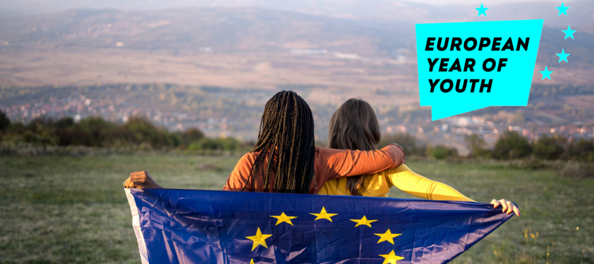 Zwei junge Frauen halten eine Europafahne über ihre Schulten und schauen über eine hügelige Landschaft