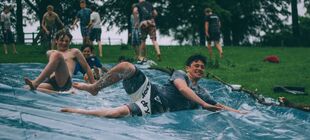 Eine Gruppe männlicher Jugendlicher rutscht auf blauen Plastikbahnen durch Seifenwasser einen Wiesenhügel herunter.