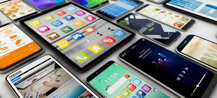 Zahlreiche Tablets und Smartphones liegen nebeneinander auf einem Tisch, bunte Icons und Apps sind zu sehen 