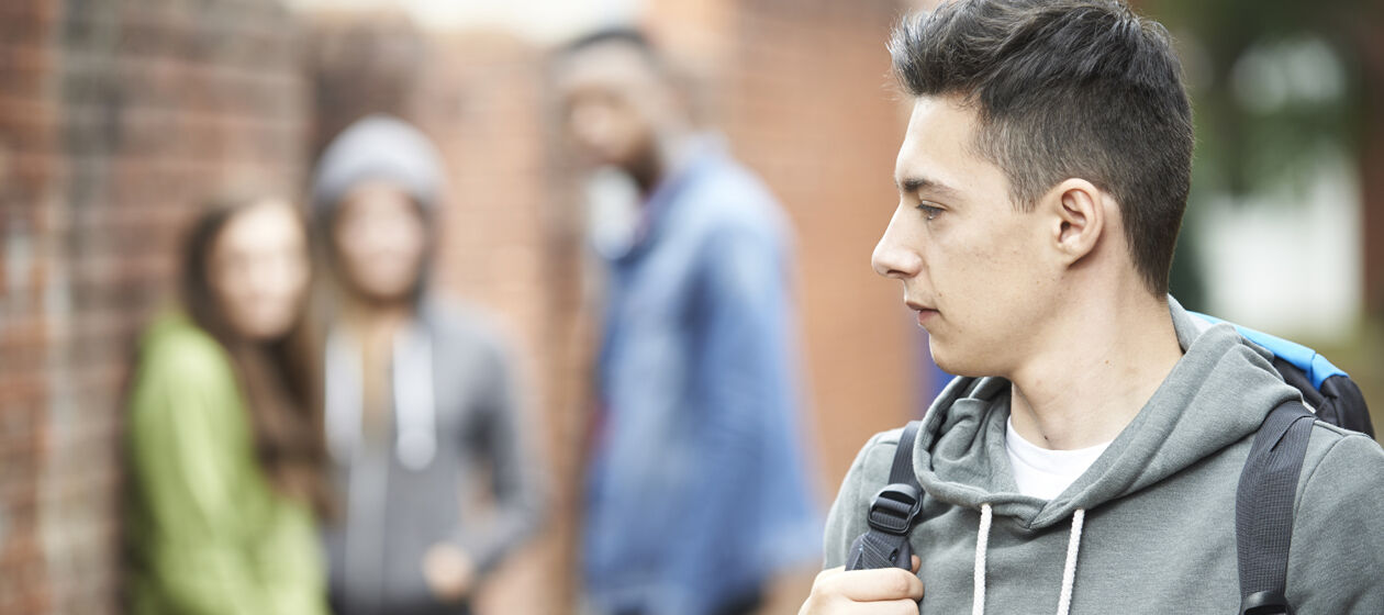 Ein Jugendlicher mit Rucksack ist im Profil zu sehen, im unscharfen Hintergrund steht eine Gruppe Jugendlicher die zu ihm schaut.