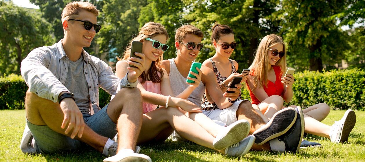 Fünf Jugendliche sitzen nebeneinander im Gras und sind auf ihr Smartphone konzentriert
