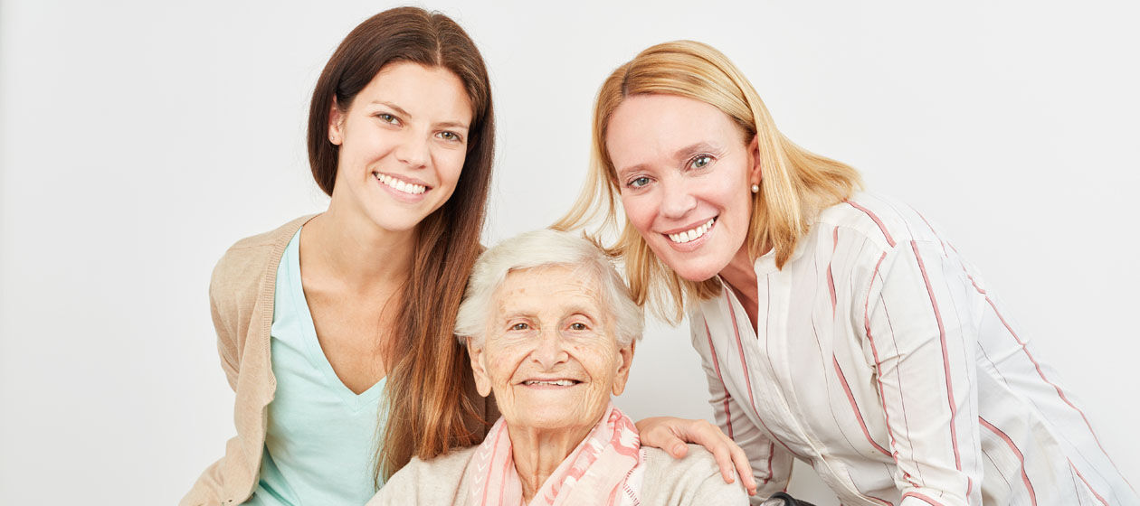 Eine Großmutter sitzt und neben ihr steht die Tochter und die Enkelin und alle drei lachen in die Kamera.