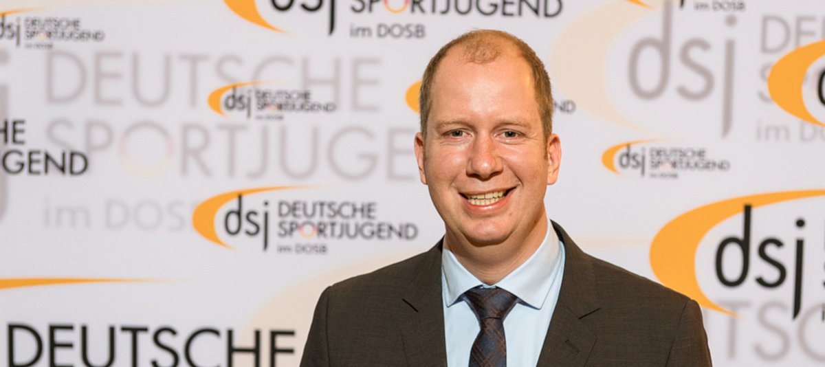 Interviewpartner Jan Holze vor einer Wand mit dem Logo der Deutschen Sportjugend