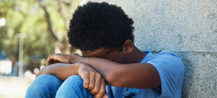 Jugendlicher afrikanischer Herkunft sitzt mit dem Kopf auf die Knie gestützt an einer Mauer auf der Strasse.