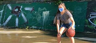 Eine Jugendliche mit Mund-Nase-Schutz spielt in einem Hof Basketball.