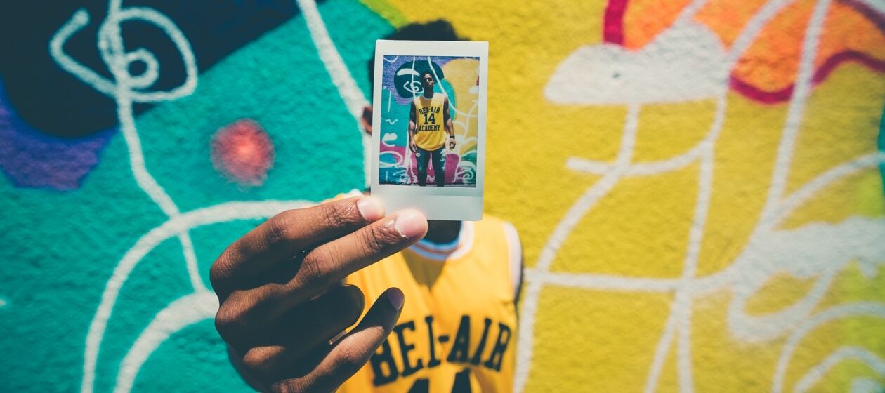 Ein Jugendlicher steht vor einer Wand mit Graffittis und hält vor seinen Kopf ein Polaroid auf dem er abgebildet ist