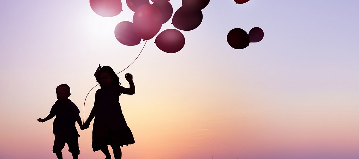 Zwei Kinder im Sonnenuntergang, die sich an der Hand halten und Luftballons steigen lassen