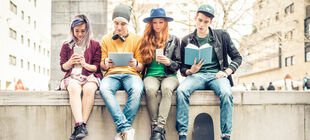 Vier Jugendliche sitzen auf einer Mauer, jeder hält ein Medium in der Hand, darunter zwei Smartphones, ein Tablet und ein Buchn 