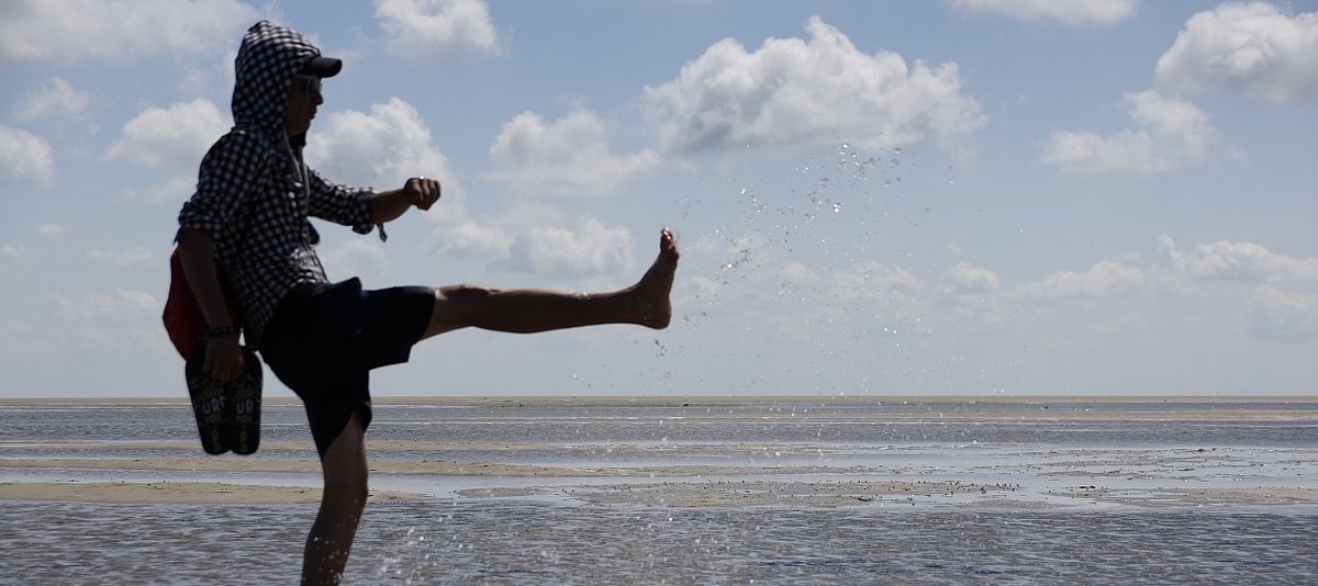 Eine junger Mann mit dunkler Hautfarbe steht am Strand und spritz energievoll mit einem Bein Wasser in die Luft